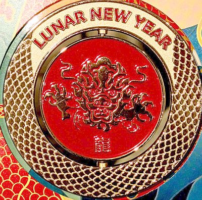 Close up of the obverse of the medallion. with LUNAR NEW YEAR in red on gold and the dragon skin pattern over the rest of the outer, and front on image of a dragon leaping towards us on the main part of the medallion, front feet either side and dragon symbol below.