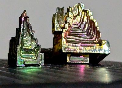 Two bismuth crystals, one green and purple and triangle shaped and one slightly squarer and stepped and green-yellow to pink-purple