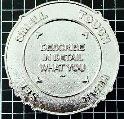 Large round coin, all plain silver coloured on this side.  In the centre, across three lines is "DESCRIBE IN DETAIL WHAT YOU" with arrows pointing to four sections on the edge, each of which corresponds to a series of bumps on the edge itself.  With one bump is "SMELL", with two bumps is "TOUCH, three bumps is HEAR and four bumps is SEE